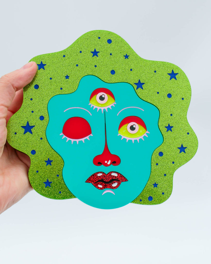 Colorful pop surrealism artwork featuring a teal three-eyed face with one eye closed, glitter hair, and red lips, art by Electric Cat.