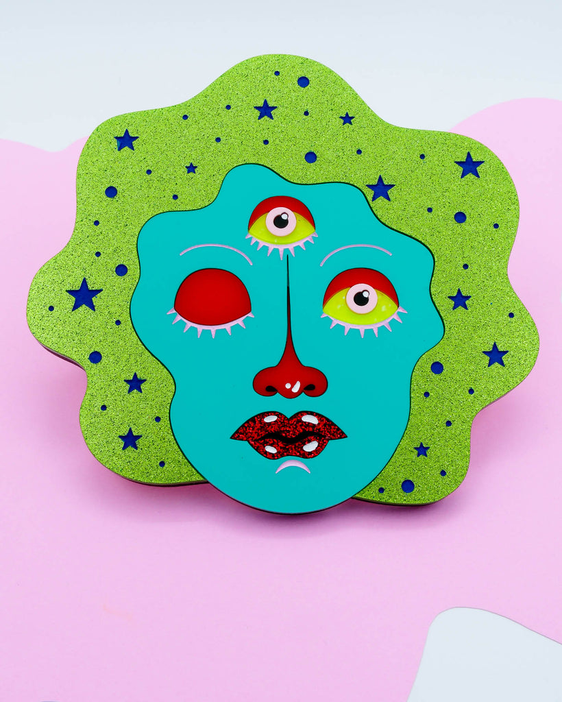 Colorful pop surrealism artwork featuring a teal three-eyed face with one eye closed, glitter hair, and red lips, art by Electric Cat.