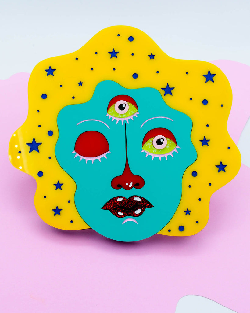 Laser-cut acrylic wall art of a three-eyed woman with yellow hair and teal face surrounded by stars, art by Electric Cat.