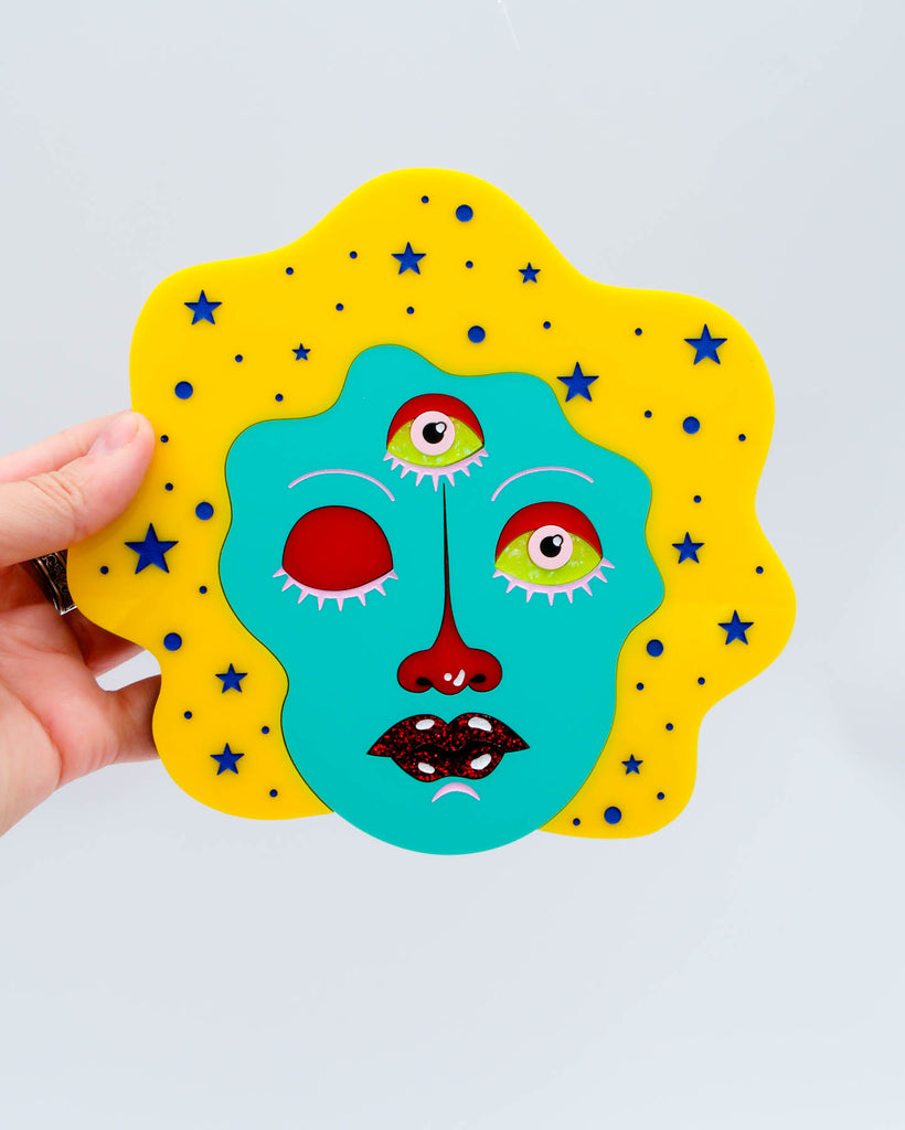 Laser-cut acrylic wall art of a three-eyed woman with yellow hair and teal face surrounded by stars, art by Electric Cat.