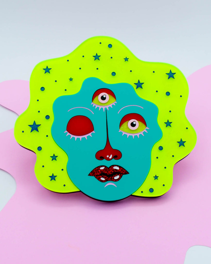 Colorful wall art of a three-eyed fortune teller face with teal skin, neon yellow hair, and glitter red lips surrounded by stars, art by Electric Cat.