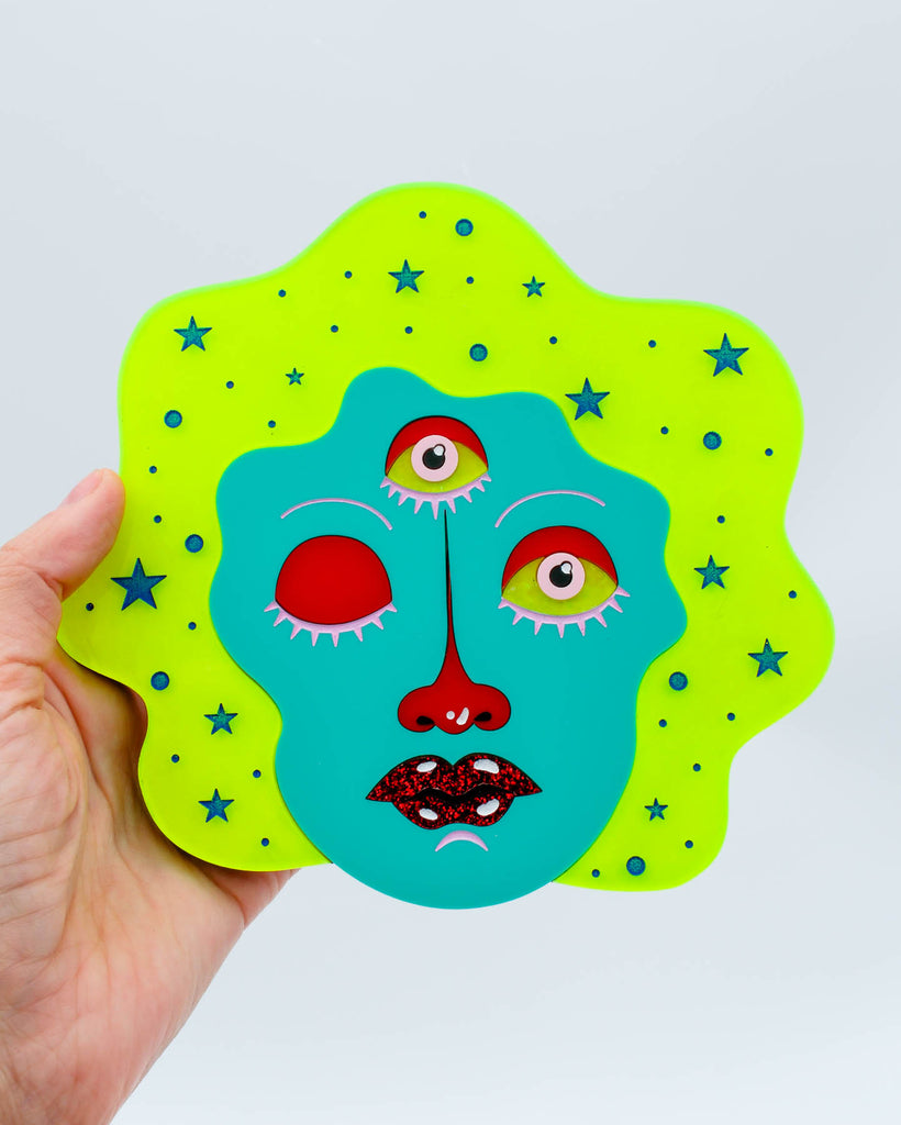 Colorful wall art of a three-eyed fortune teller face with teal skin, neon yellow hair, and glitter red lips surrounded by stars, art by Electric Cat.