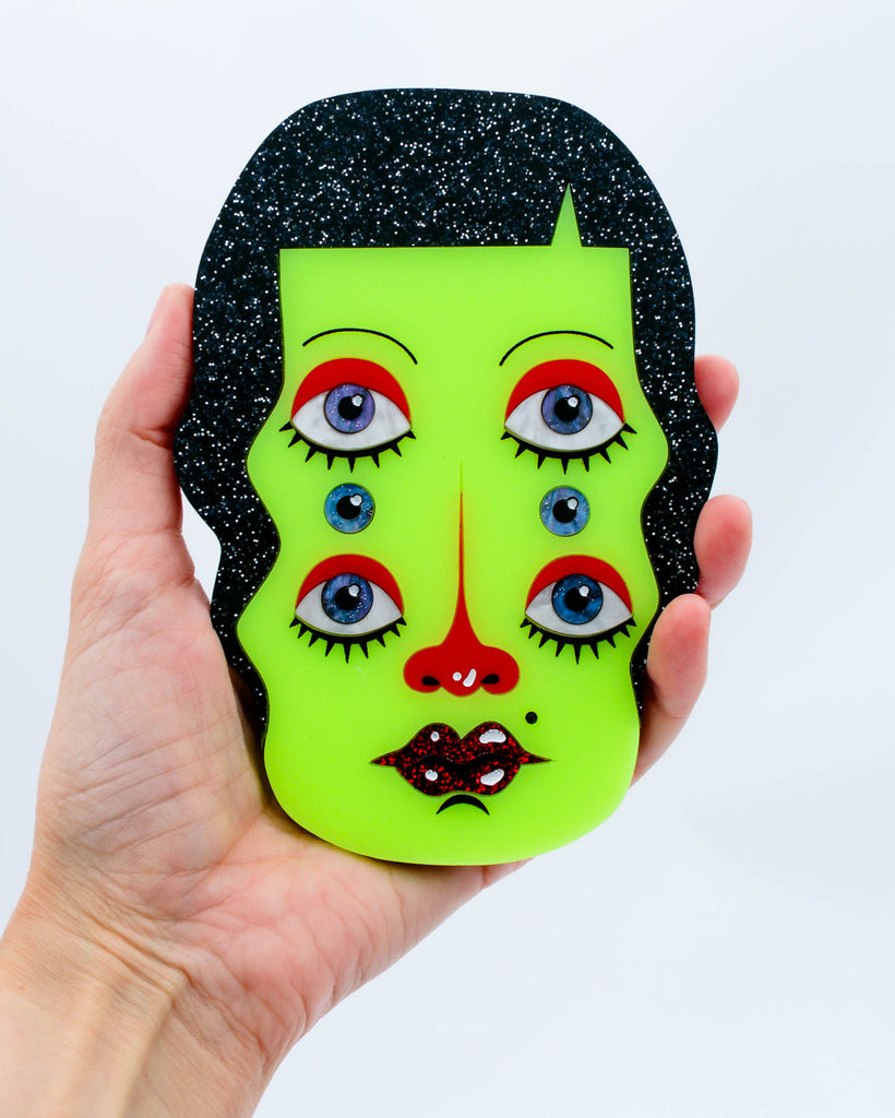 Wall art of a surreal green six-eyed woman with black glitter hair and red glitter lips, art by Electric Cat.
