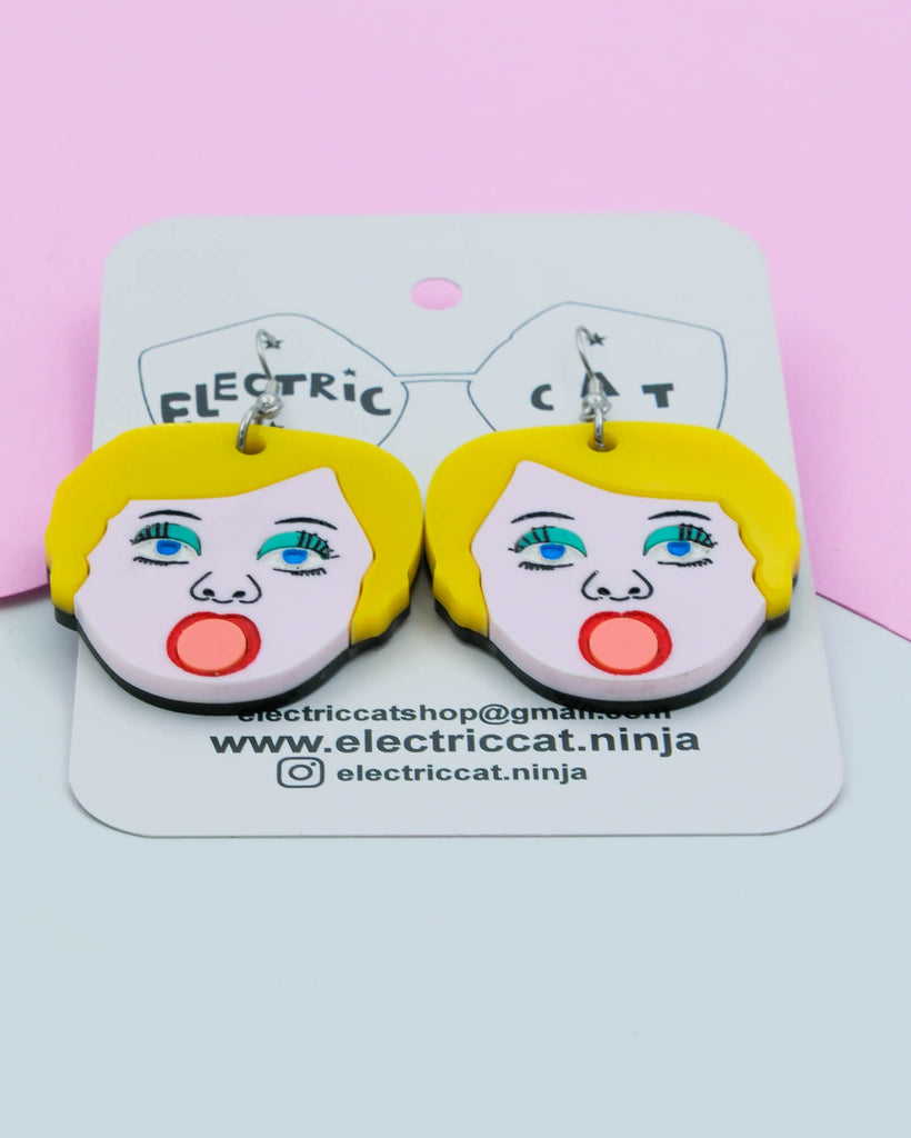 Handmade acrylic earrings shaped like blow up doll faces with yellow hair, pink lips, and teal eye shadow, art by Electric Cat.