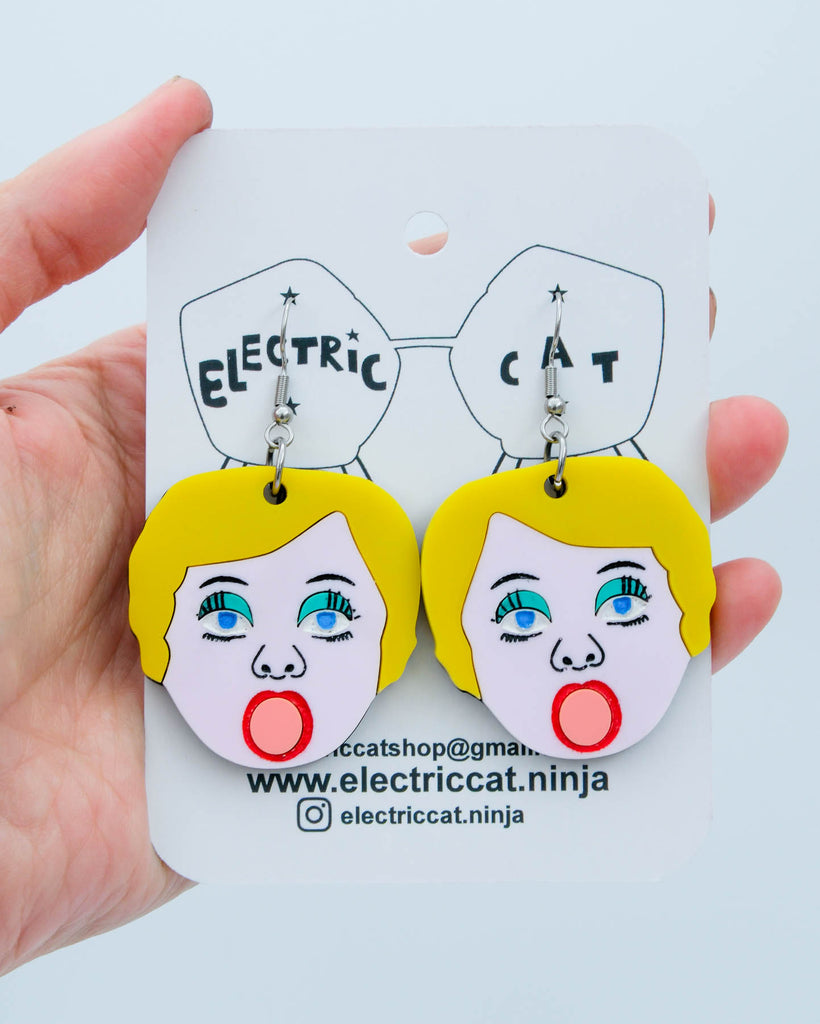 Handmade acrylic earrings shaped like blow up doll faces with yellow hair, pink lips, and teal eye shadow, art by Electric Cat.
