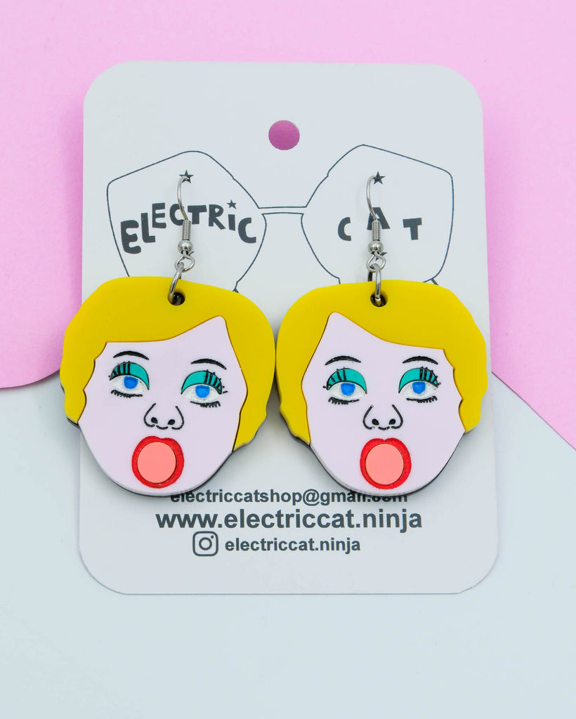 Handmade acrylic earrings shaped like blow up doll faces with yellow hair, pink lips, and teal eye shadow, art by Electric Cat.