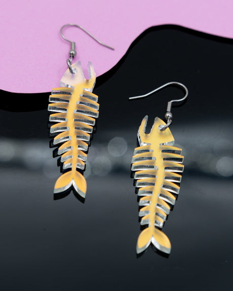 A pair of iridescent acrylic fish skeleton earrings with stainless steel hooks, displayed against a pink and black background.