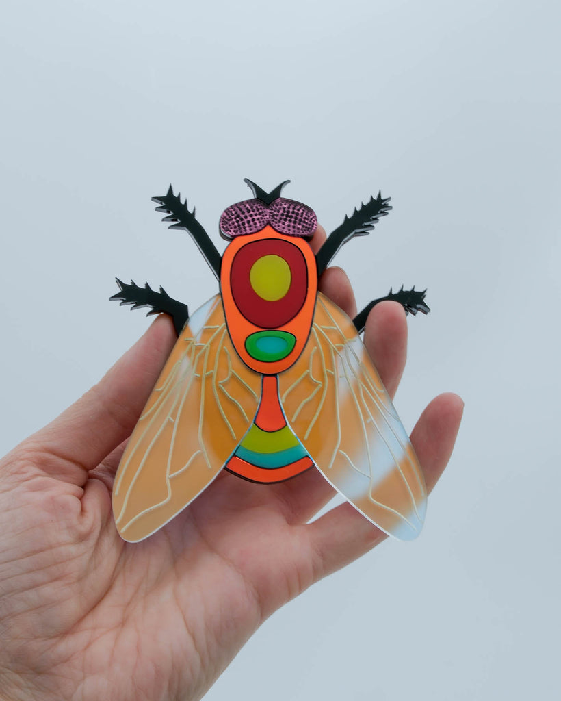 Handmade acrylic fly art piece featuring iridescent wings, black legs, and layered colorful body with pink mirrored eyes, crafted by Electric Cat.