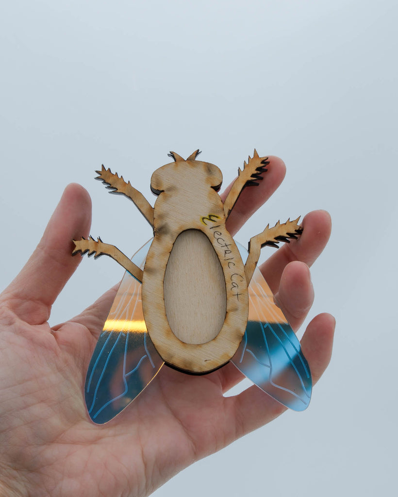 Handmade acrylic fly art piece featuring iridescent wings, black legs, and layered colorful body with pink mirrored eyes, crafted by Electric Cat.