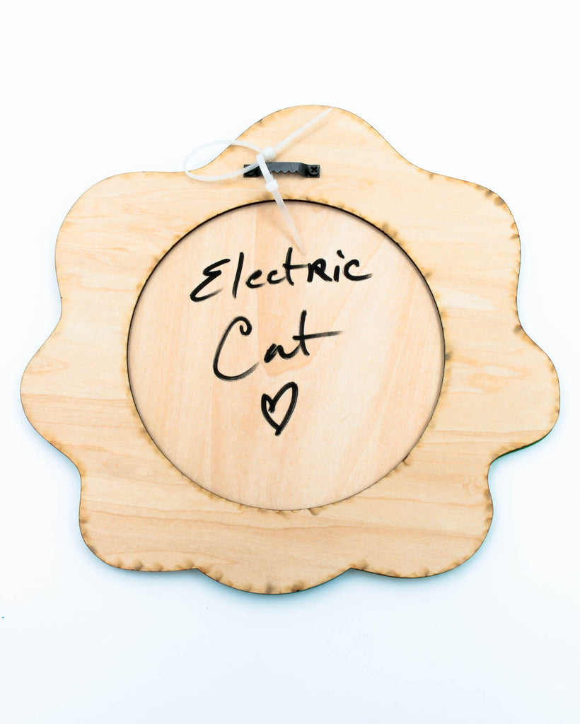 Wood back of the colorful pop surrealism wall art featuring a teal three-eyed face with one eye closed and sparkling green background, art by Electric Cat.