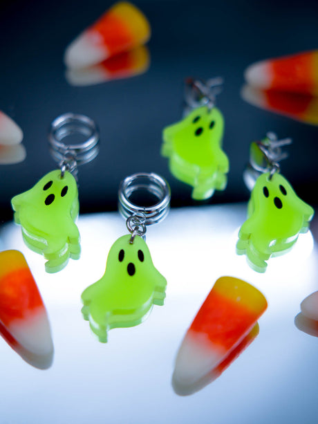 Four neon green ghost earrings with black eyes and silver metal hoops and studs, displayed on a reflective surface with candy corn props.