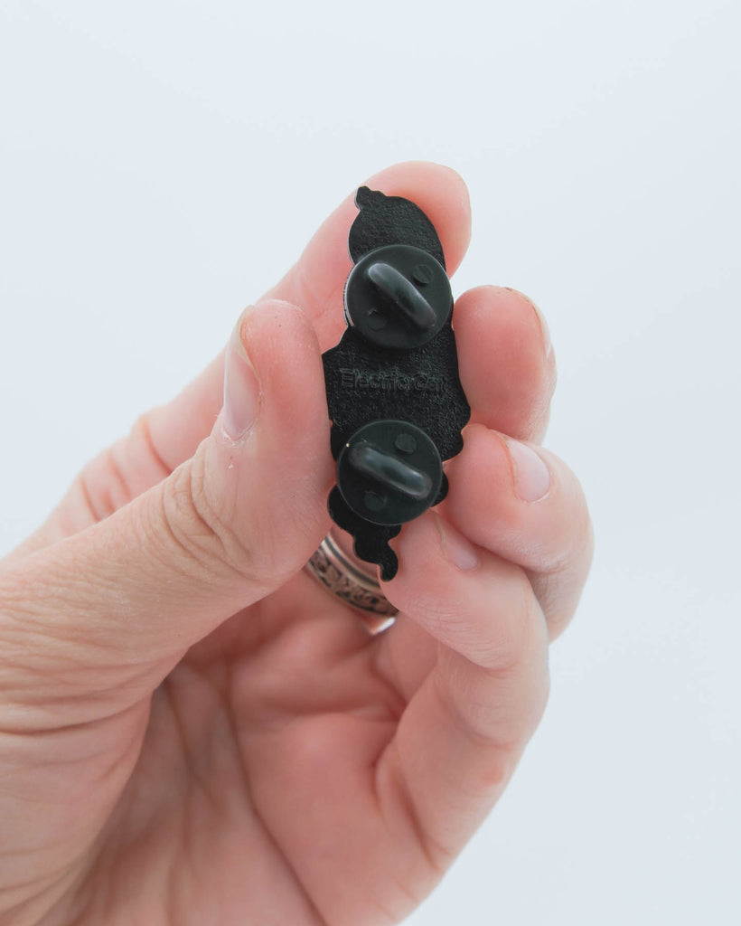 Hand holding a cartoon ghost enamel pin showing it's back against a white background