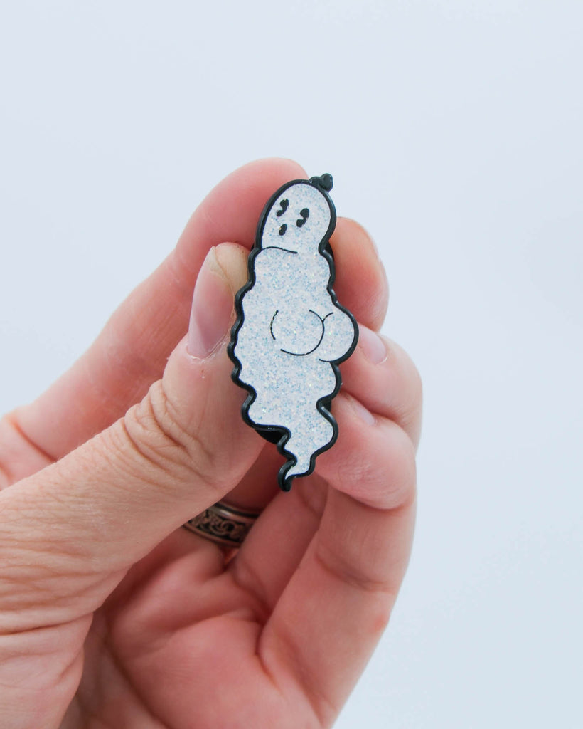 Hand holding a cartoon ghost enamel pin against a white background