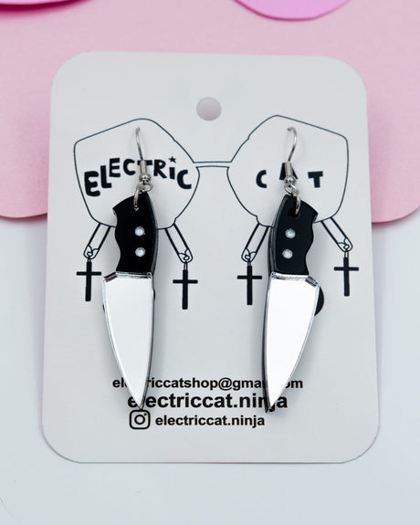 A pair of silver acrylic knife earrings with hand painted details, displayed on a pink background with 'Electric Cat' branding.