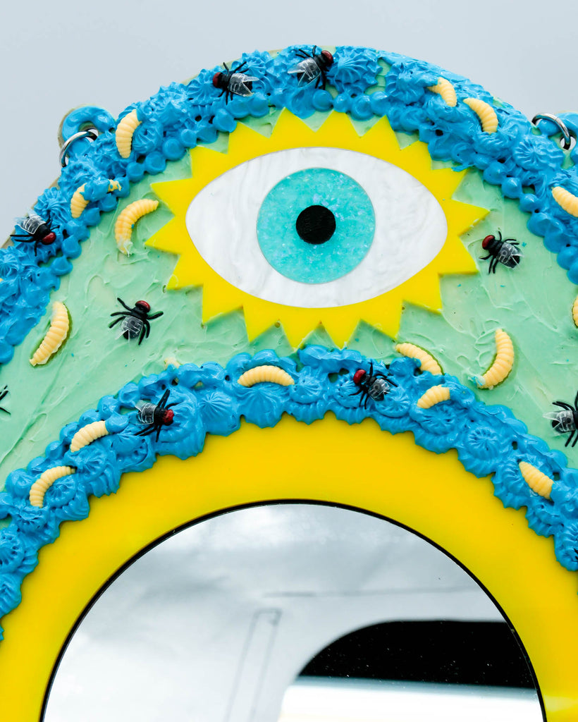 Surreal handmade mirror with textured faux frosting, two glitter eyes, flies, and maggots on blue and yellow frame, art by Electric Cat.
