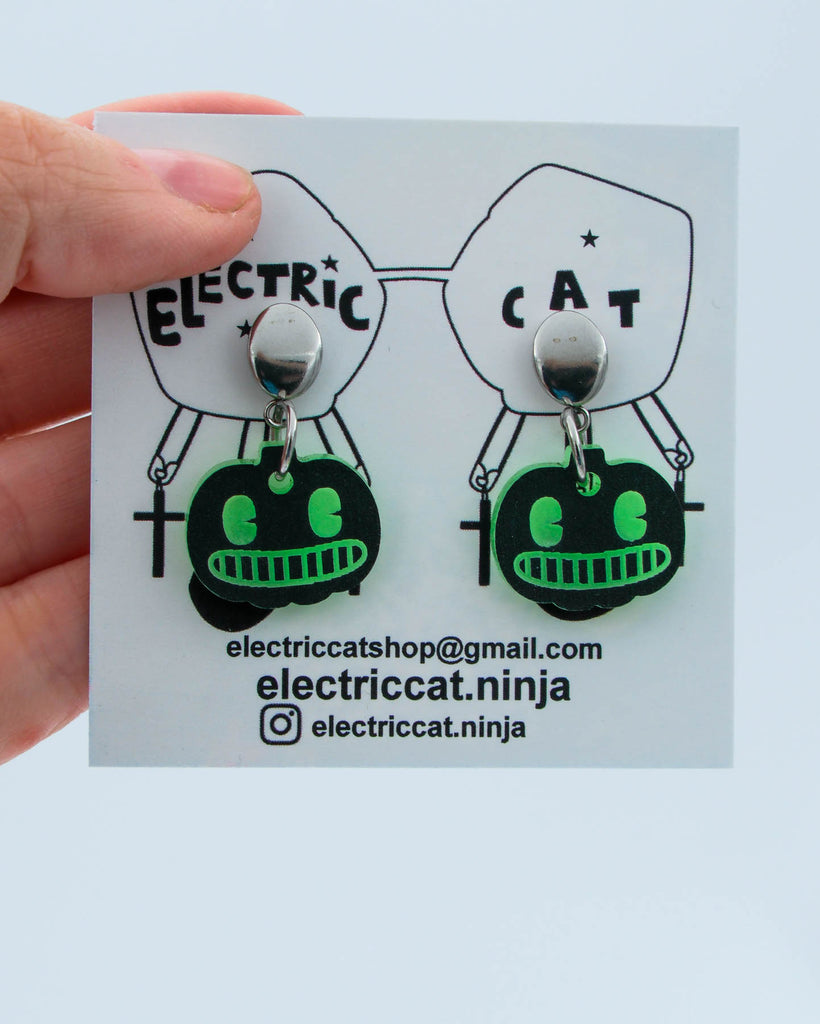 Pair of green and black pumpkin earrings on a branded card held by a hand.