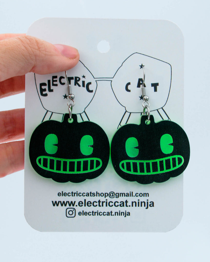 Black and green pumpkin earrings on a branded card held by a hand.