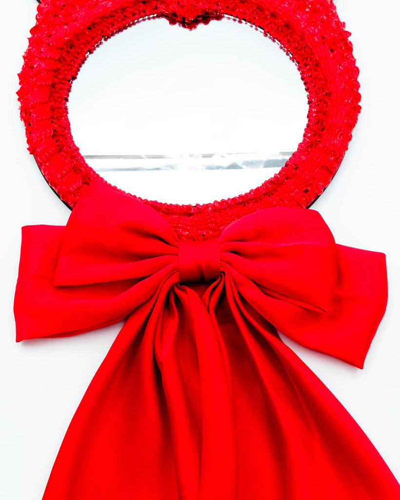 Red devil-themed mirror with sculpted frosting details, horns, and long satin ribbon bow, art by Electric Cat.