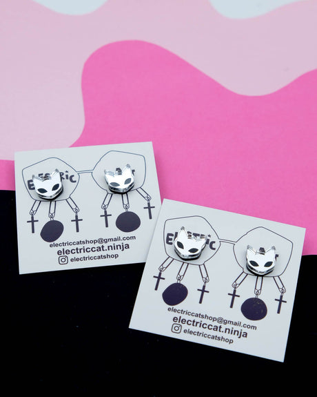 A pair of acrylic cat earrings with a silver mirrored finish, laid on a black surface with a pink background.